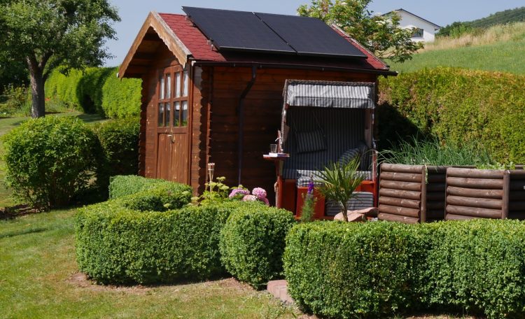 Holzhütte mit Solarpanel, umgeben von gepflegtem Grün und Hecken.